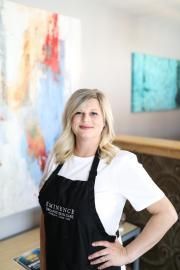 Woman in a black apron standing in an art studio with colorful paintings on the wall