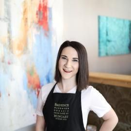 Smiling woman in a black apron standing in a bright studio beside colorful abstract art