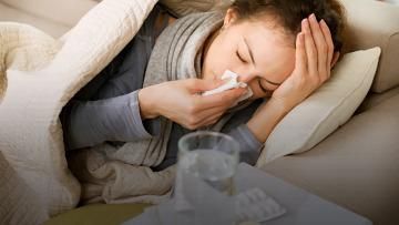 Person lying in bed, blowing their nose with a tissue beside a glass of water