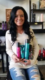 Woman sitting indoors holding several hair care products, smiling in front of salon shelves.