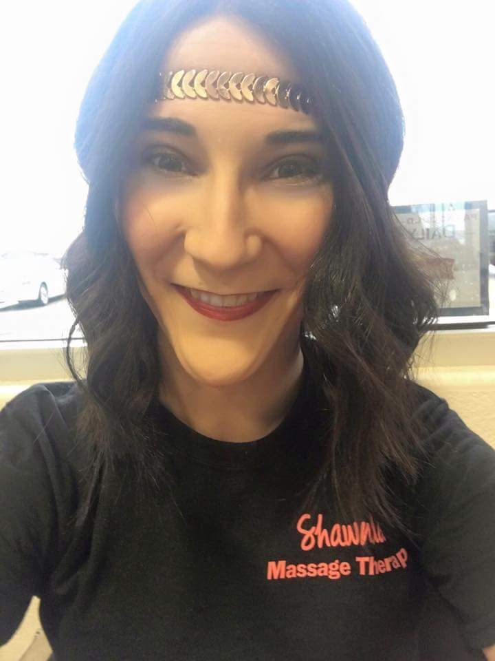 Smiling woman in black shirt with red “Massage Therapist” text, standing indoors by a window.