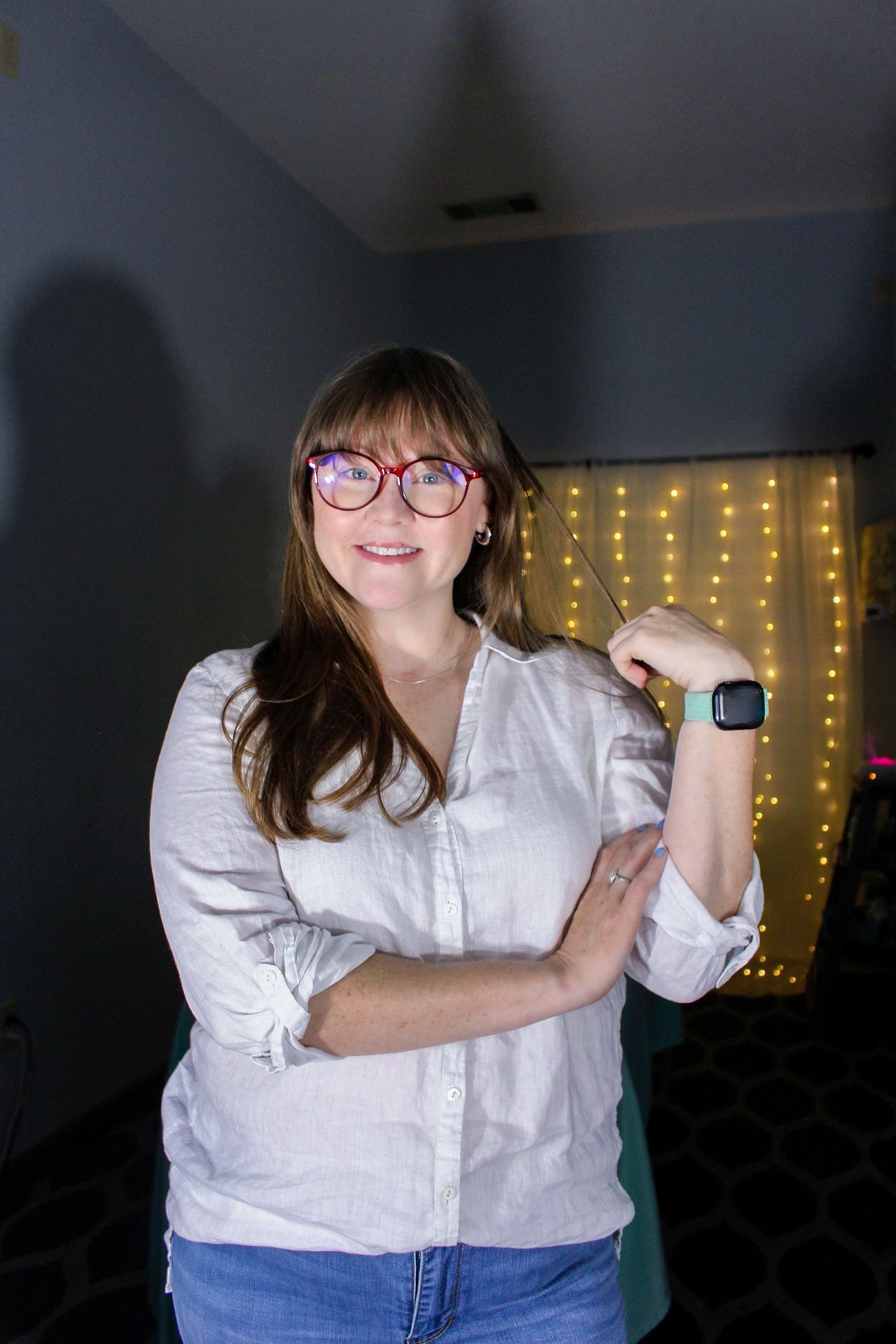 Person in glasses and white shirt posing indoors with a smartwatch, in front of string lights.