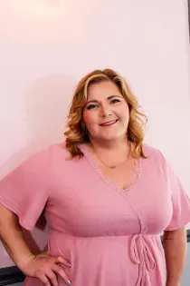 Smiling person in a pink dress, standing with hand on hip against a light pink wall.