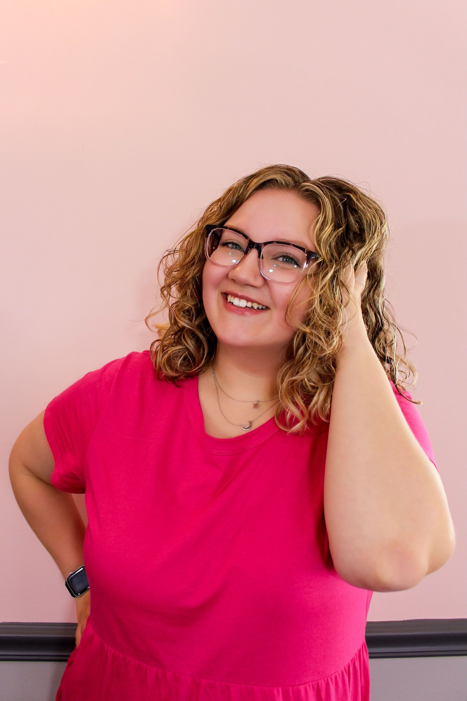 Smiling person with curly hair and glasses in a bright pink shirt, standing against a pale pink wall.