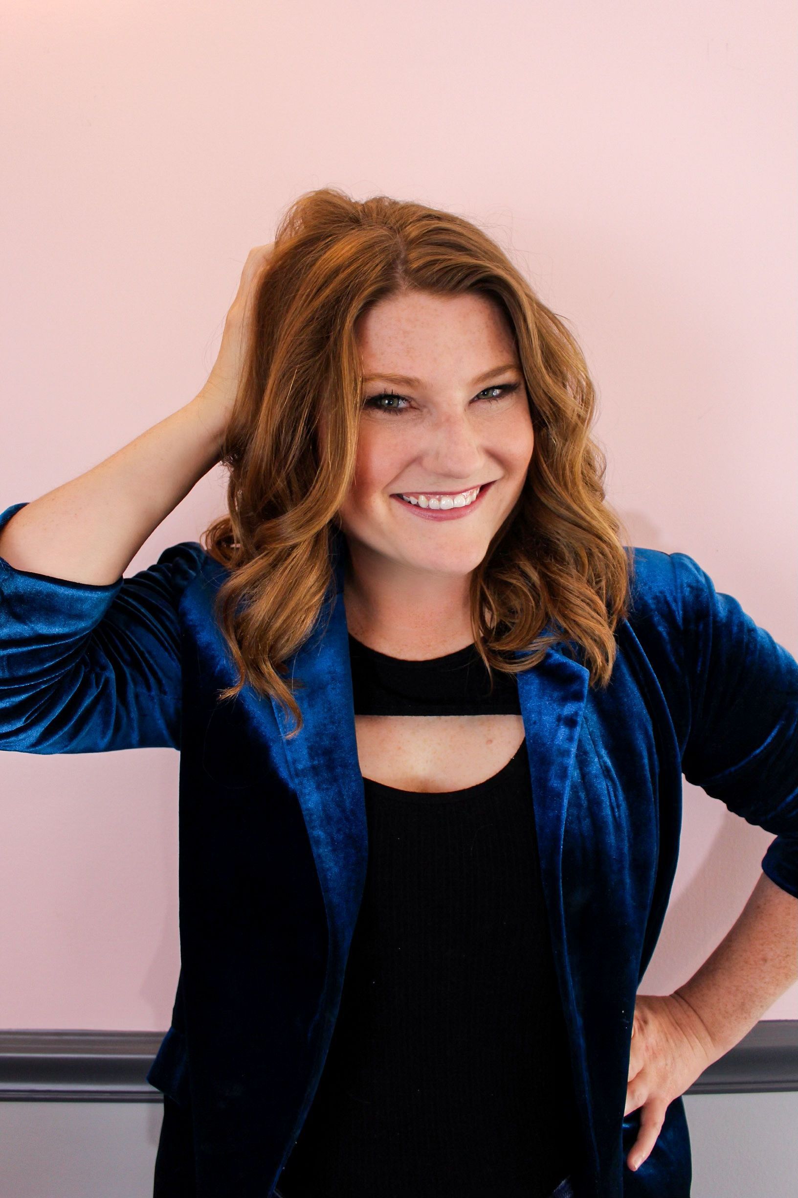 Smiling woman in a blue jacket, posing with one hand on her head against a pink wall.
