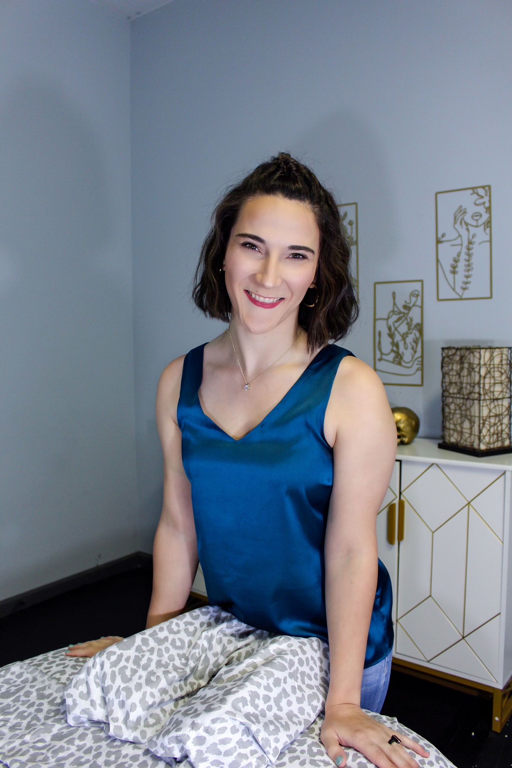 Woman sitting on a bed in a blue top, smiling in a softly lit bedroom.