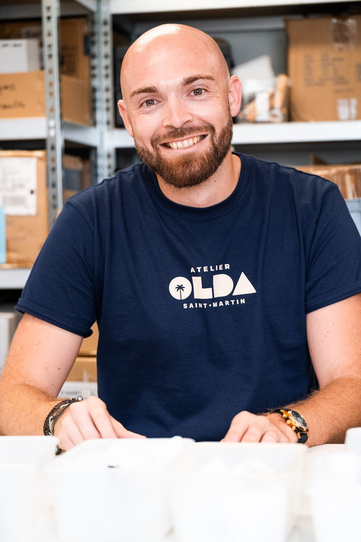 Un homme chauve avec une barbe portant un t-shirt bleu est assis à une table dans un entrepôt.