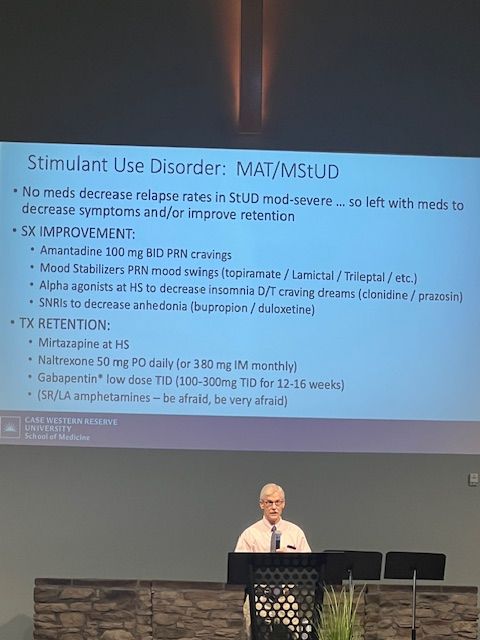 Conference slide about “Stimulant Use Disorder” projected above a speaker at a podium on a lecture stage