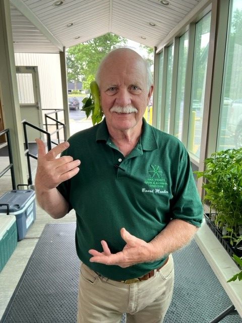 Man in green polo shirt smiling and gesturing on a covered porch with windows and plants