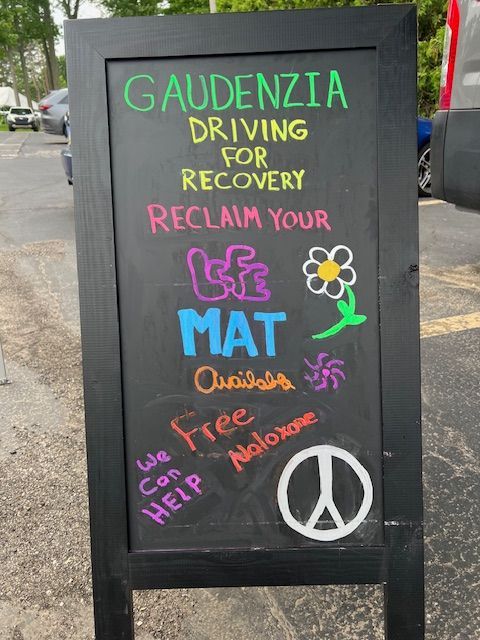 Handmade sidewalk sign reading “Gaudenzia Driving or Recovery, Reclaim Your MAT,” with colorful drawings and a peace symbol.