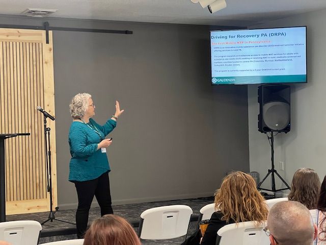 Presenter in teal jacket speaks beside a projected slide in a small meeting room.