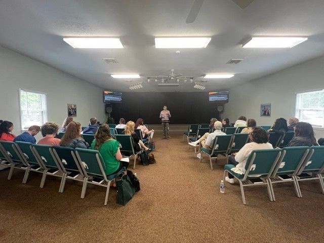 Audience seated in a meeting room facing a speaker at the front of the room.
