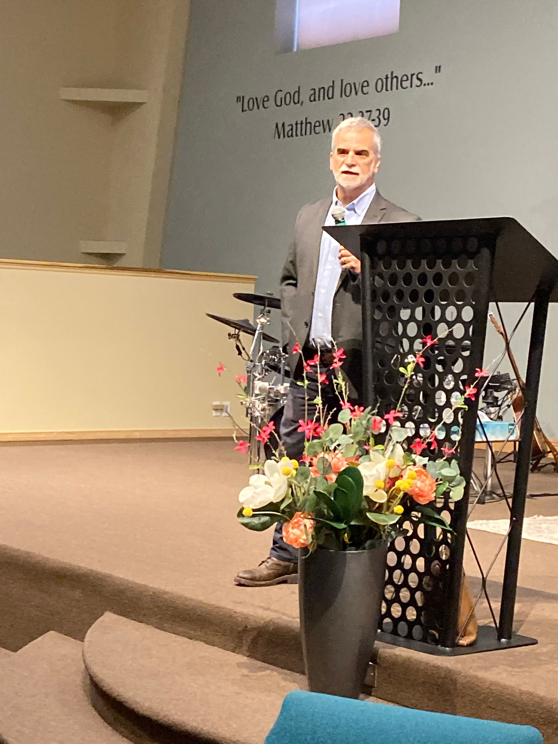Man speaking at a church lectern with flowers in front and steps leading to the stage.