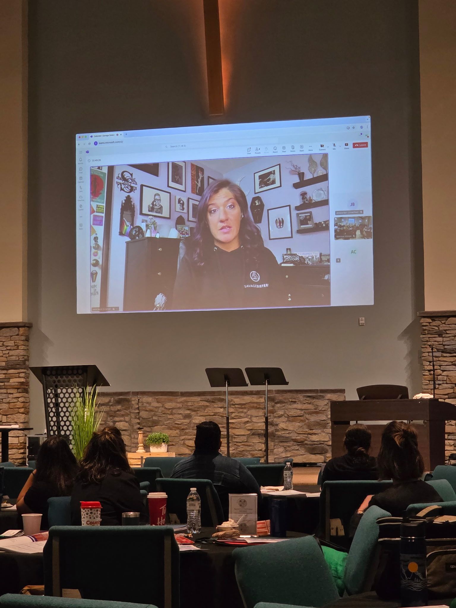 Audience watching a projected video call in a dim auditorium with rows of seats and a stone stage backdrop