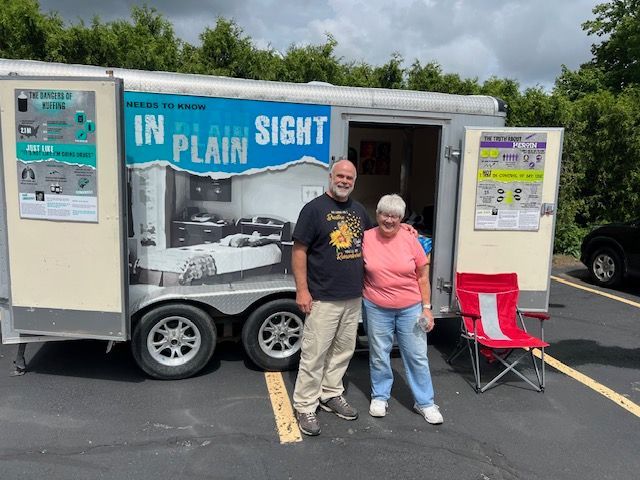 Two people standing beside a mobile exhibit trailer labeled “In Plain Sight” at an outdoor event