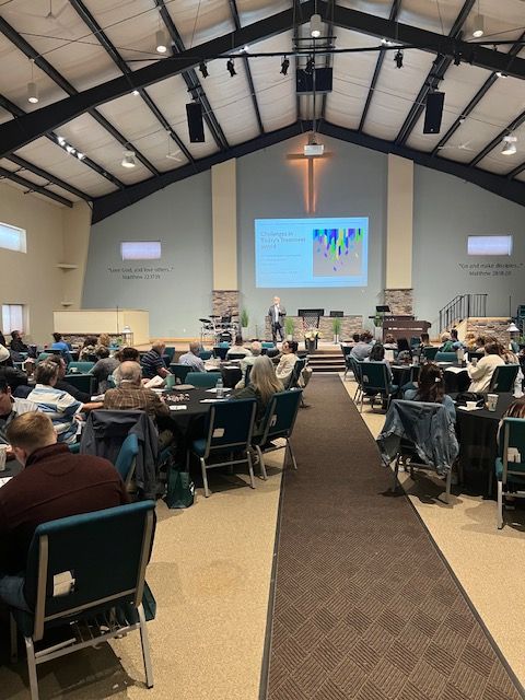 Large church hall with people seated at tables facing a speaker and projection screen during a presentation.