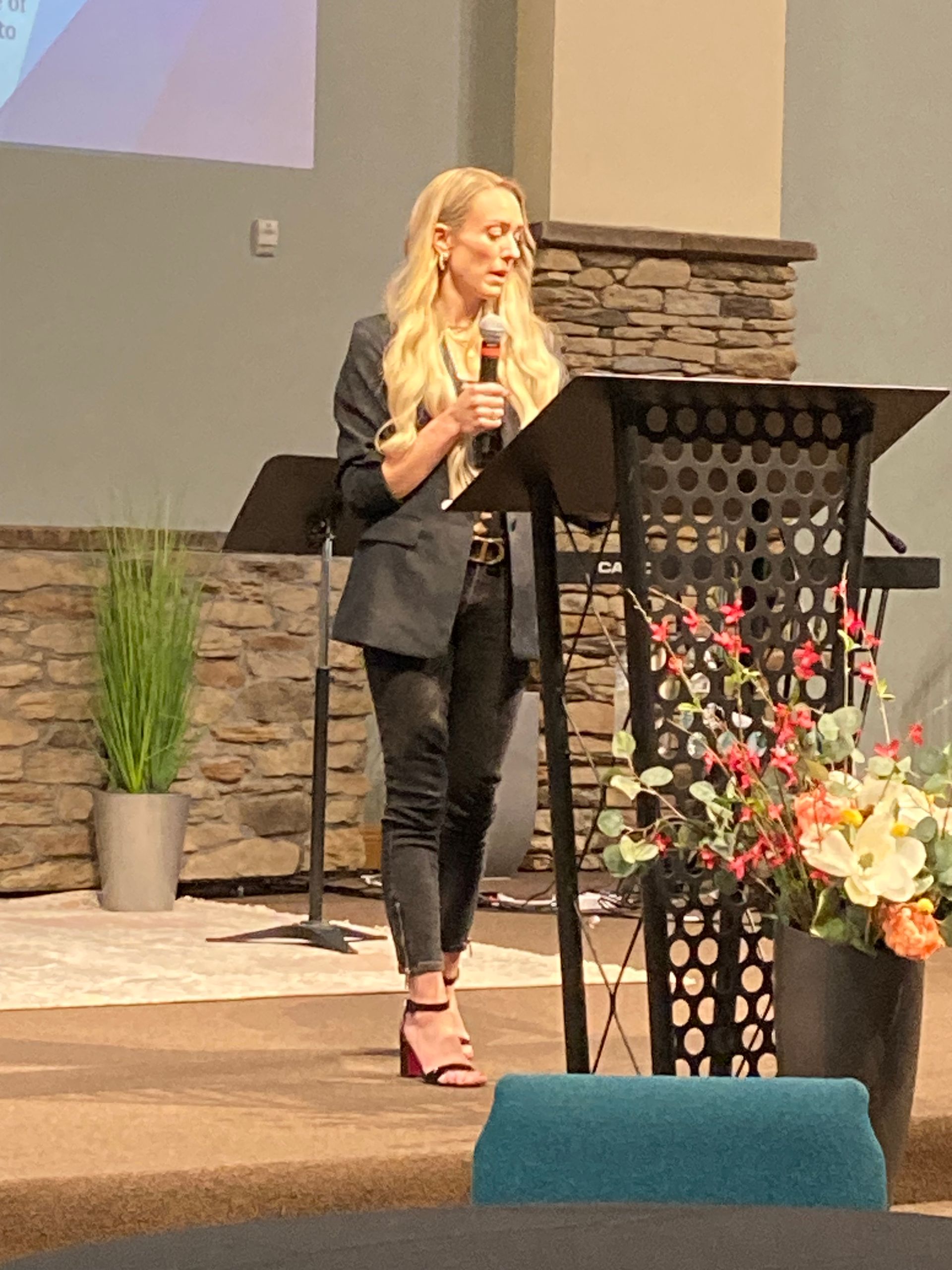 Woman speaking at a church podium beside flowers, holding a microphone on a stage.