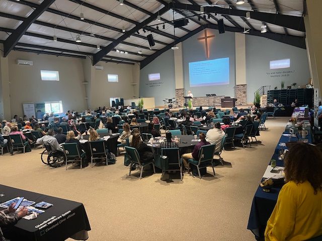 Large hall with many people seated at tables for a conference or banquet, facing a projected screen and cross on the wall
