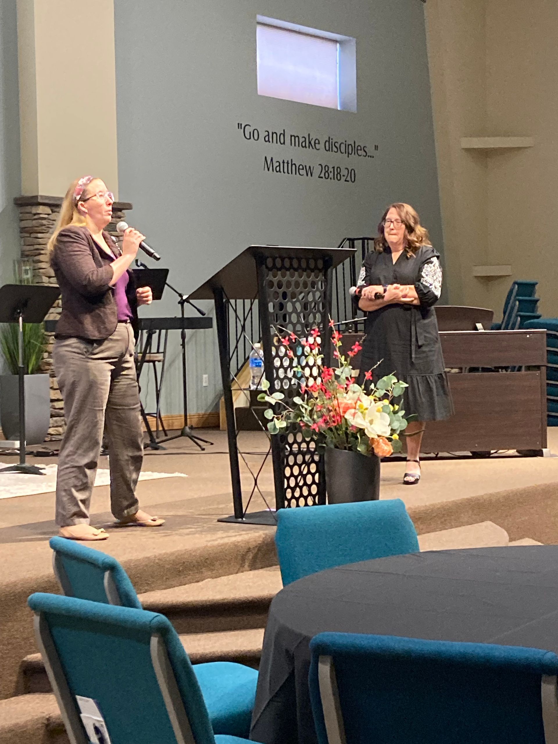 Two women speaking on a church stage beside a lectern and floral arrangement