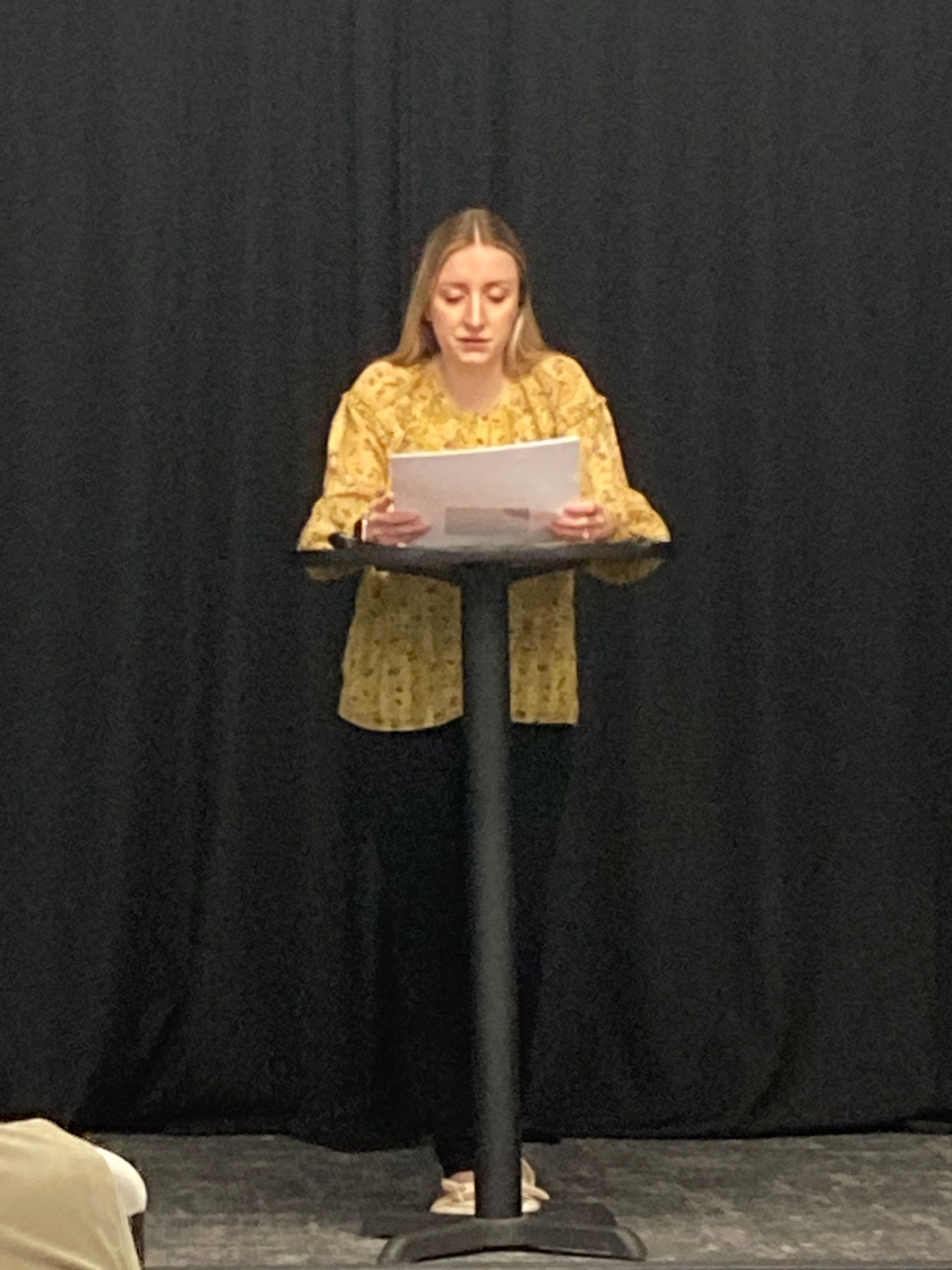 Person reading from papers at a podium on a stage with black curtains