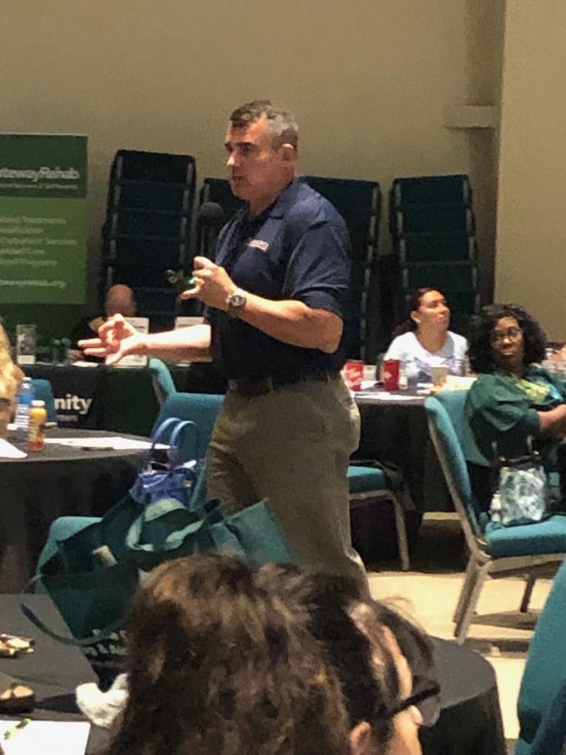 Man speaking at a conference, holding a microphone in a room with seated attendees.