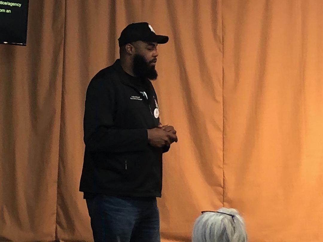 Man in a black cap and hoodie stands onstage before orange curtains, speaking to an audience.