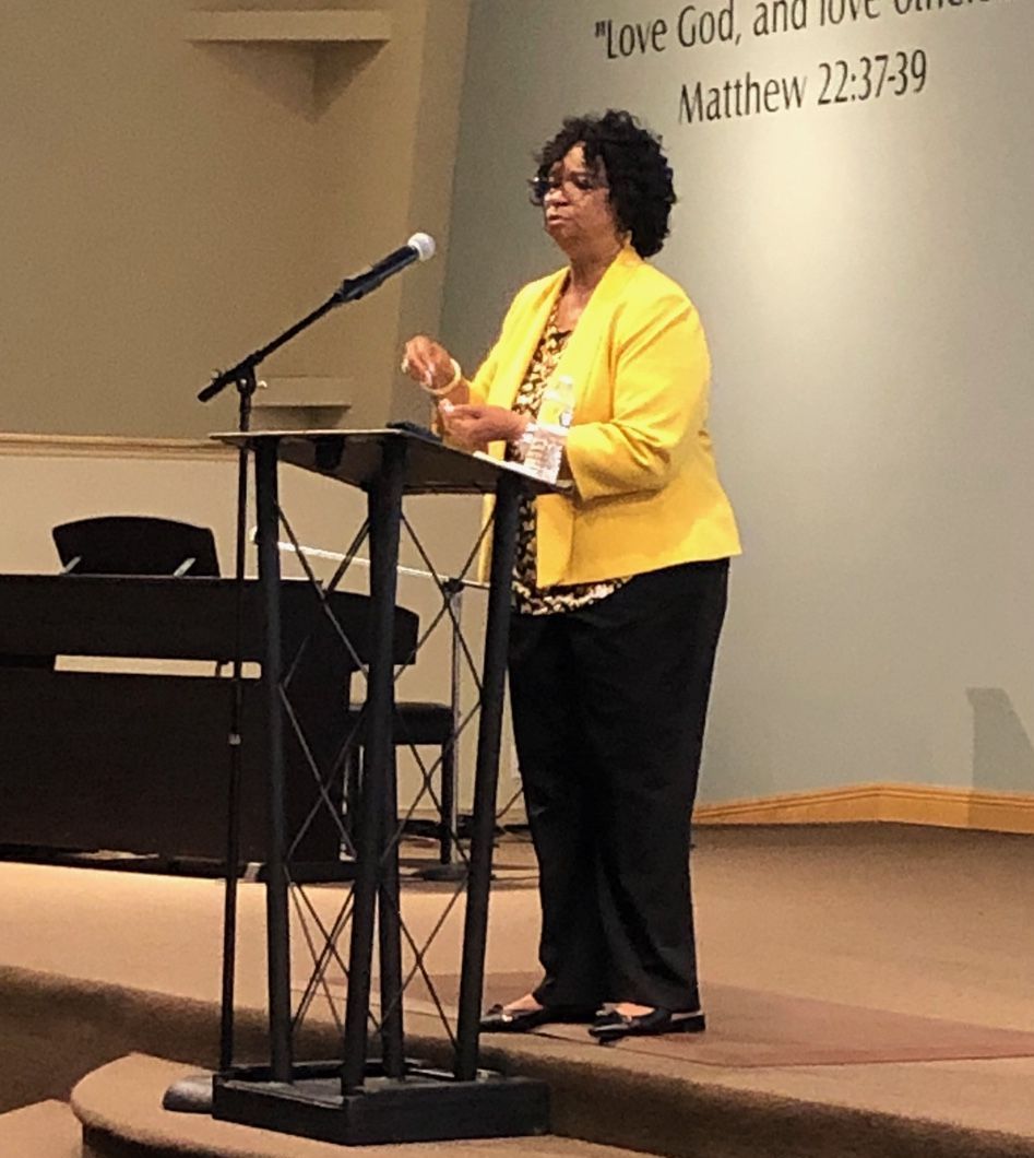 Woman speaking at a church podium, wearing a yellow jacket beside a microphone and lectern.