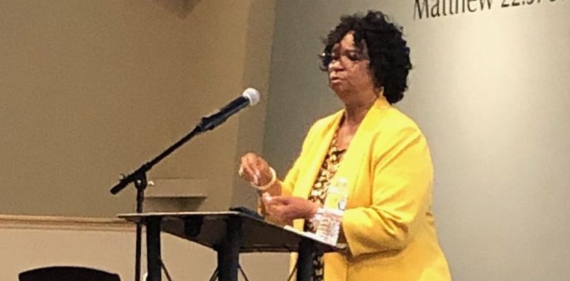 Speaker in a yellow jacket stands at a podium beside a microphone, gesturing during a presentation.