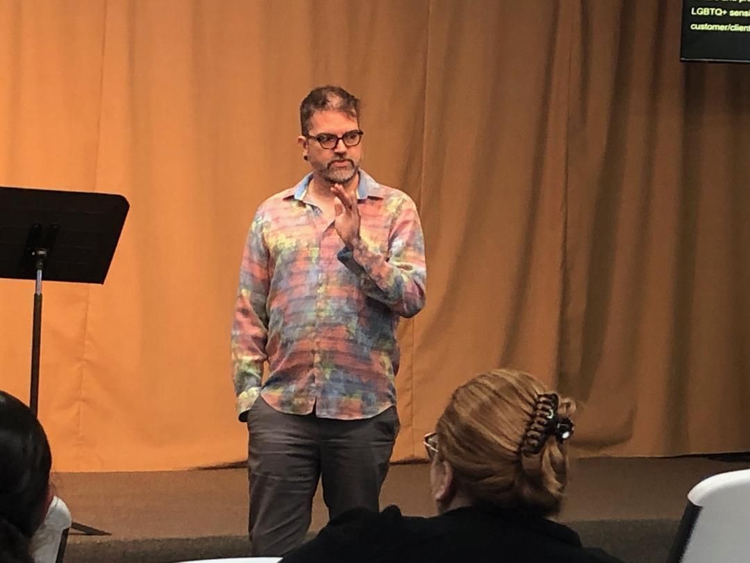 Man speaking onstage with a microphone, wearing a colorful shirt, in front of a tan curtain.