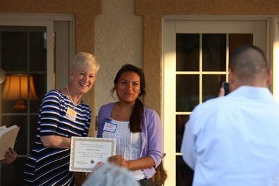 Two women posing with a certificate on a porch, with a man seen from behind in the foreground.