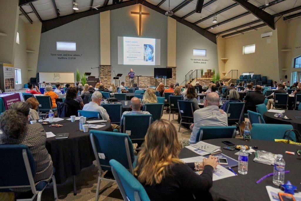 Conference audience seated in a church hall facing a presentation screen and cross at the front.