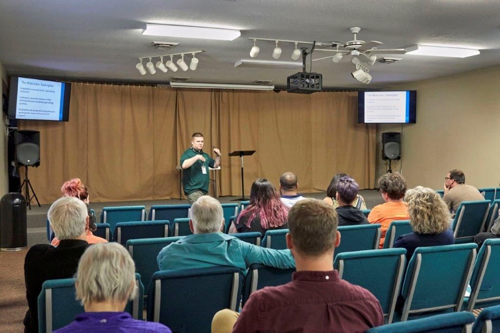 Audience seated in a small hall watching a speaker at the front, with two screens on either side.