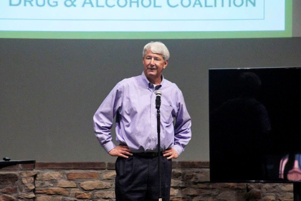 Man in a light purple shirt speaking at a microphone on a stage with a DRUG & ALCOHOL COALITION banner behind him