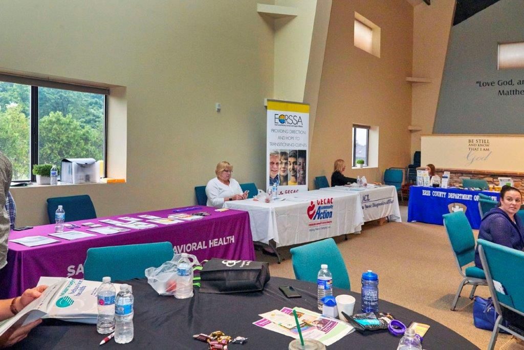 Conference room with vendor tables, banners, and seated attendees at a community event.