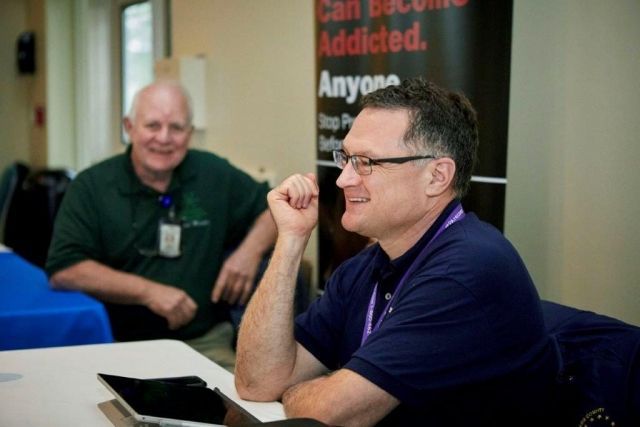 Two men sitting at a table in a meeting room, one smiling and the other listening with a tablet nearby.