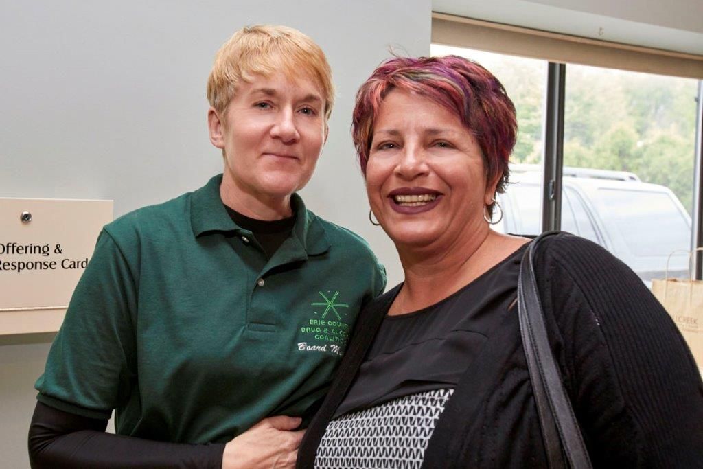 Two people smiling indoors, one in a green polo and one in a black top, standing near a window.