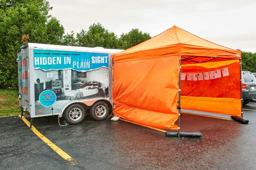 Orange canopy tent beside a “Hidden in Plain Sight” display trailer in a wet parking lot