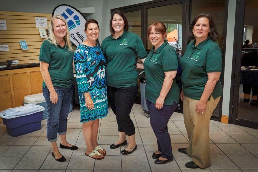 Five people in green shirts pose indoors, smiling in front of a tiled wall.