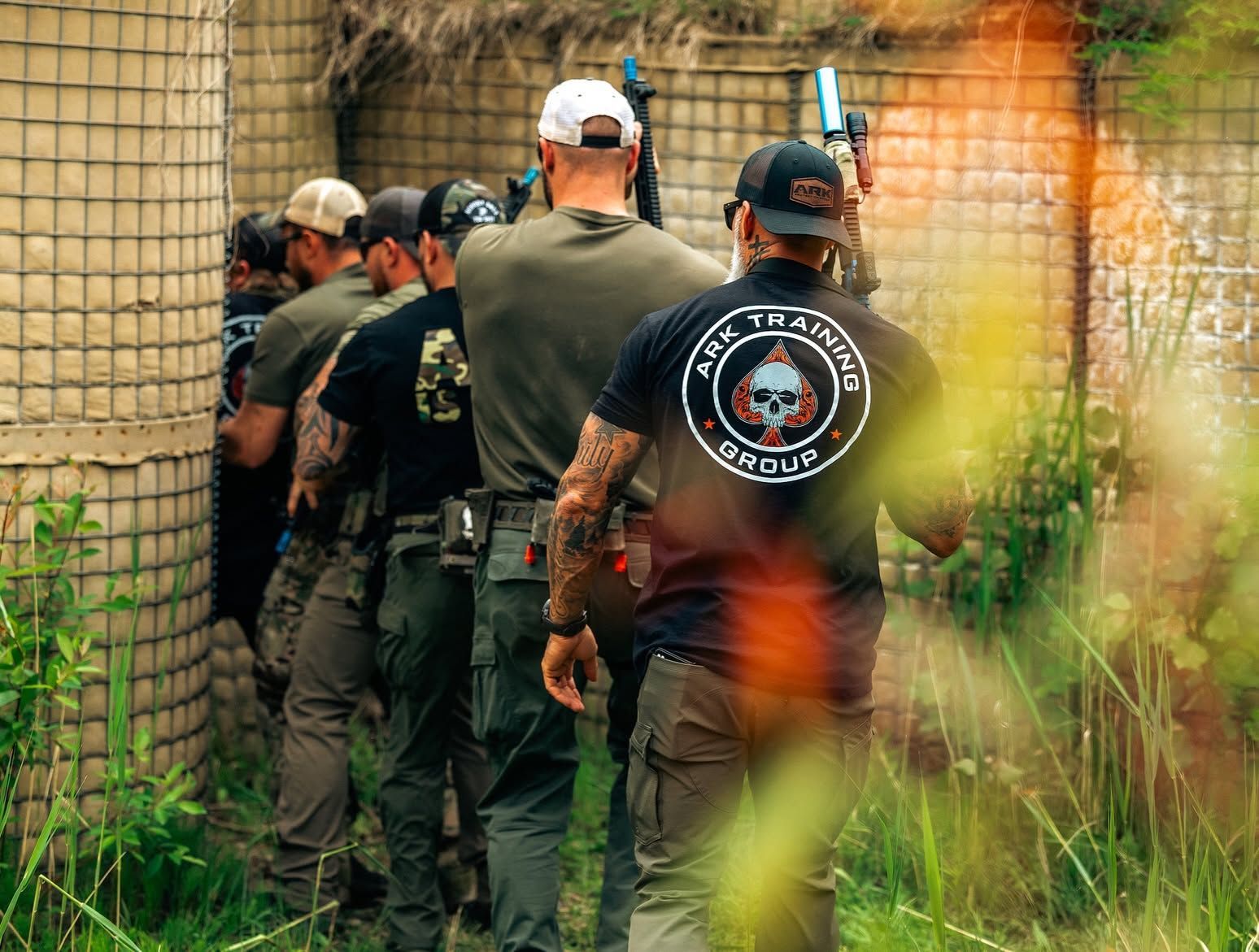 Group of people in tactical gear walk through a narrow outdoor training lane beside gabions.