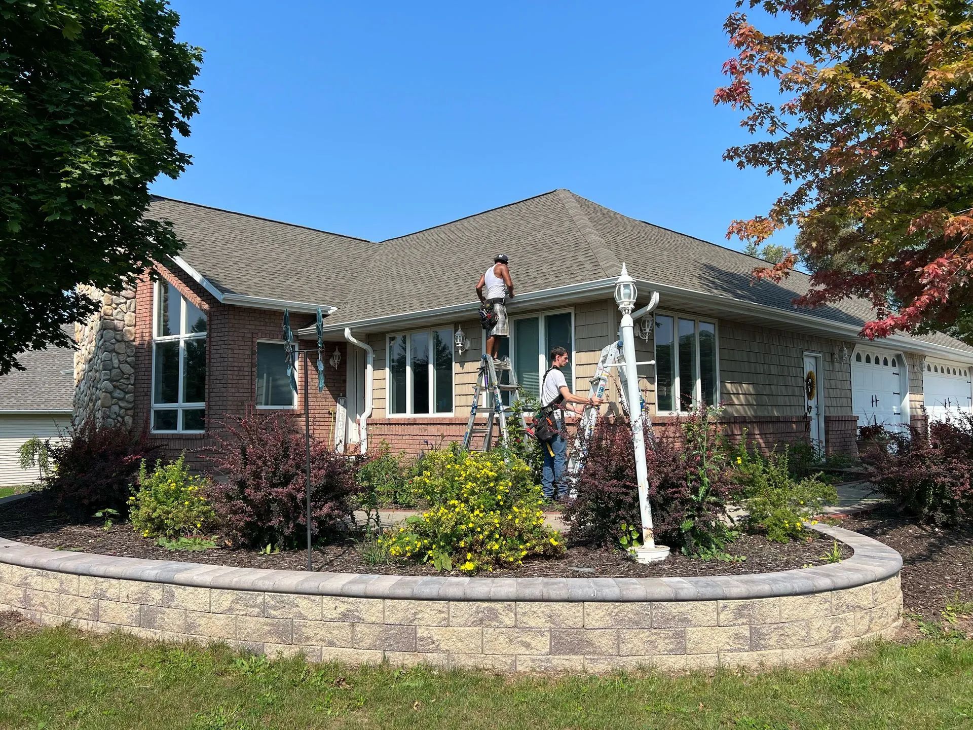 Single-story brick house with a landscaped front yard and stone retaining wall under a blue sky