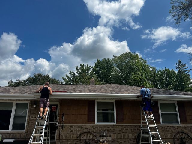 Two people stand on ladders, installing gutters on the front of a one-story house under a blue sky with clouds.