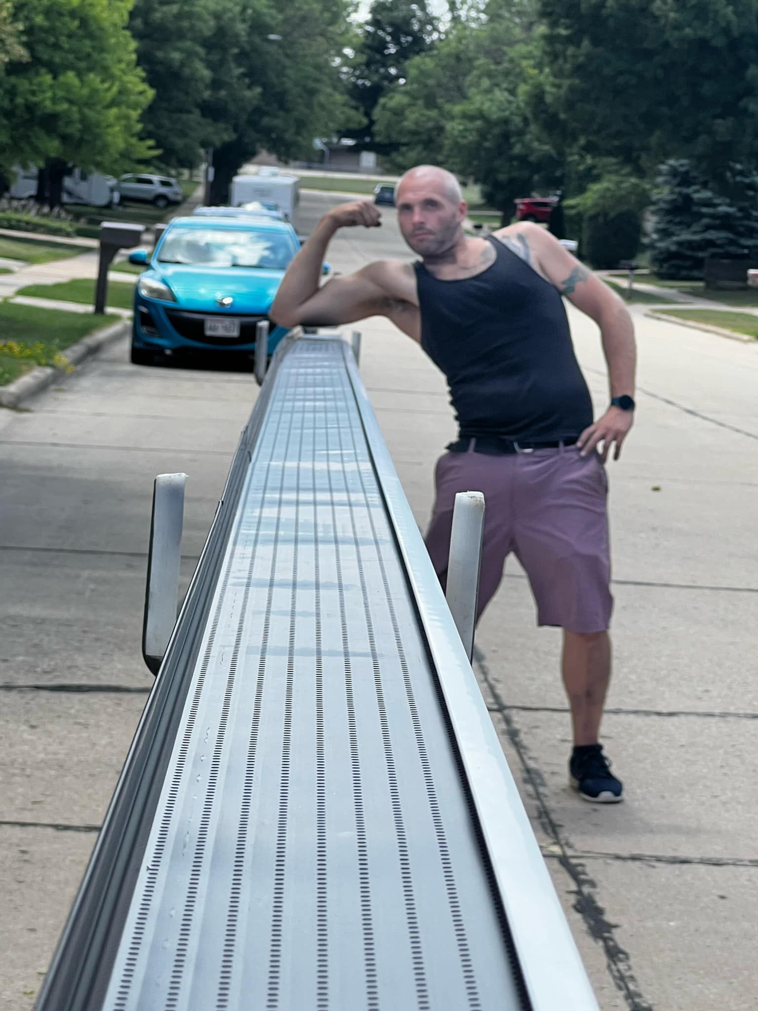A man stands on a residential street flexing his arm, leaning against a long, grey piece of metal building material.