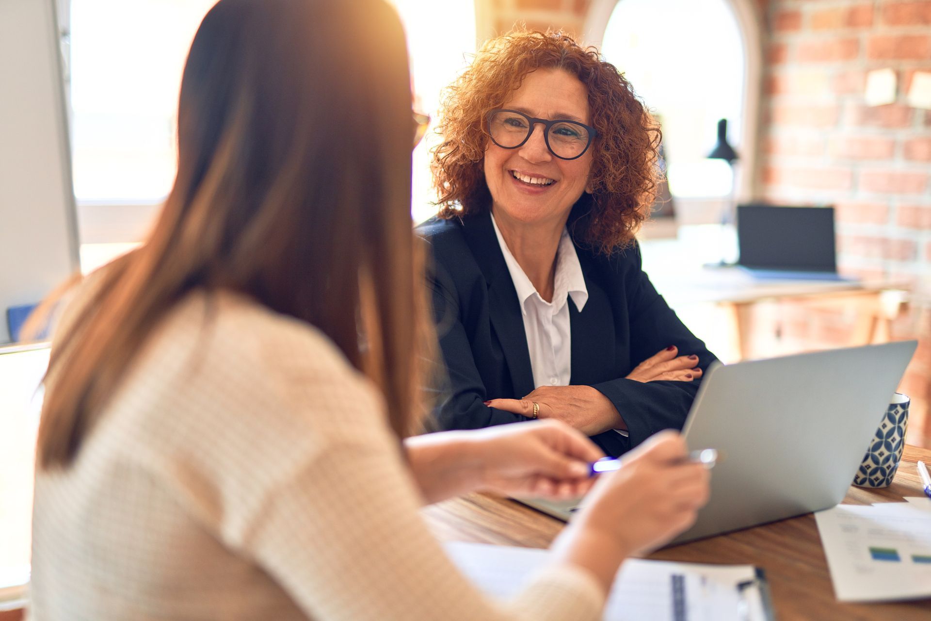 Two women at a desk talking, one smiling with one laptop open. 
