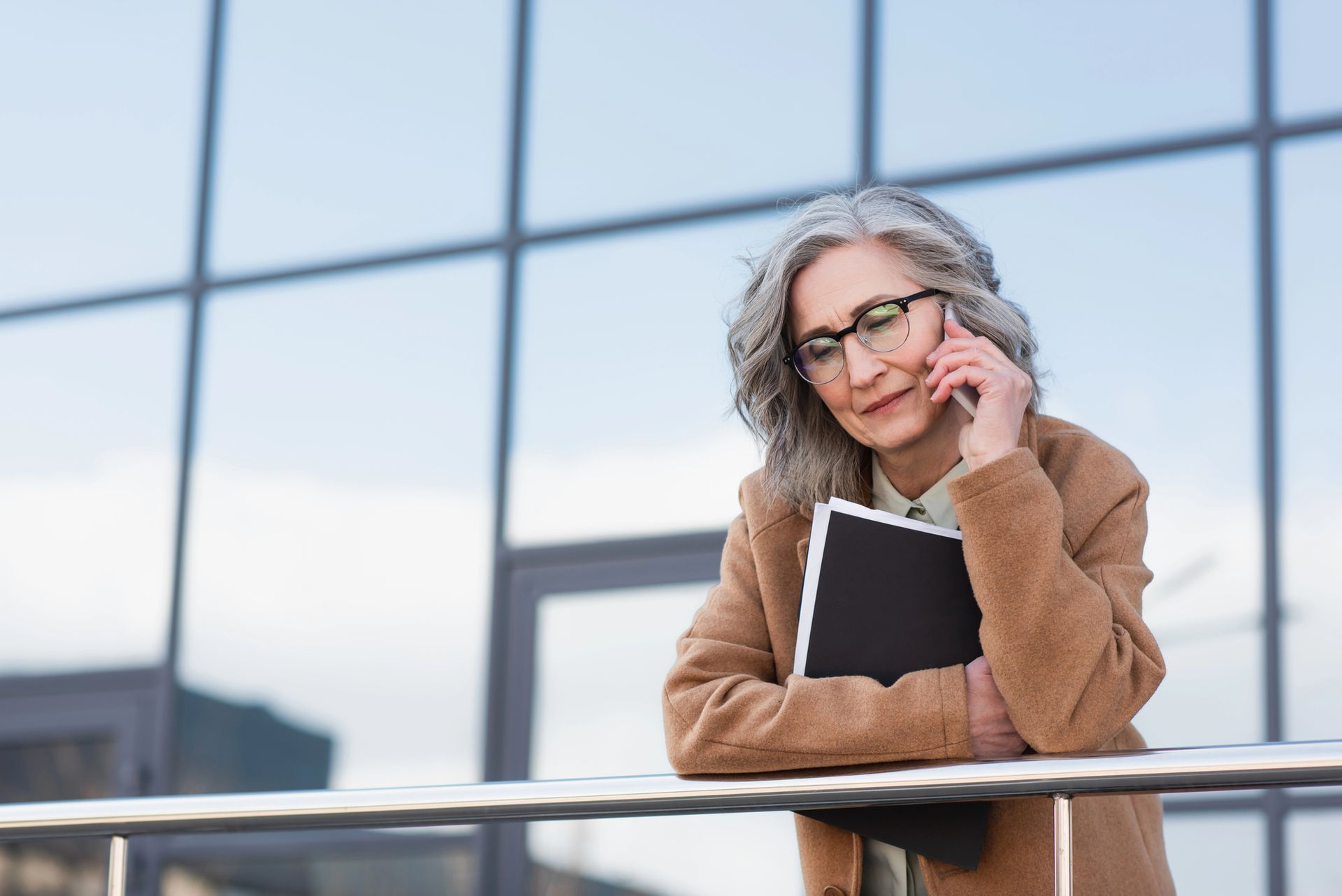 Gray haired businesswoman in a camel coat leans on a railing outside a glass office building, holding a folder while talking on her phone