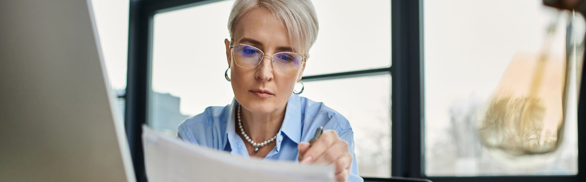 Mature woman with short silver hair wearing glasses and reading paperwork with window in background