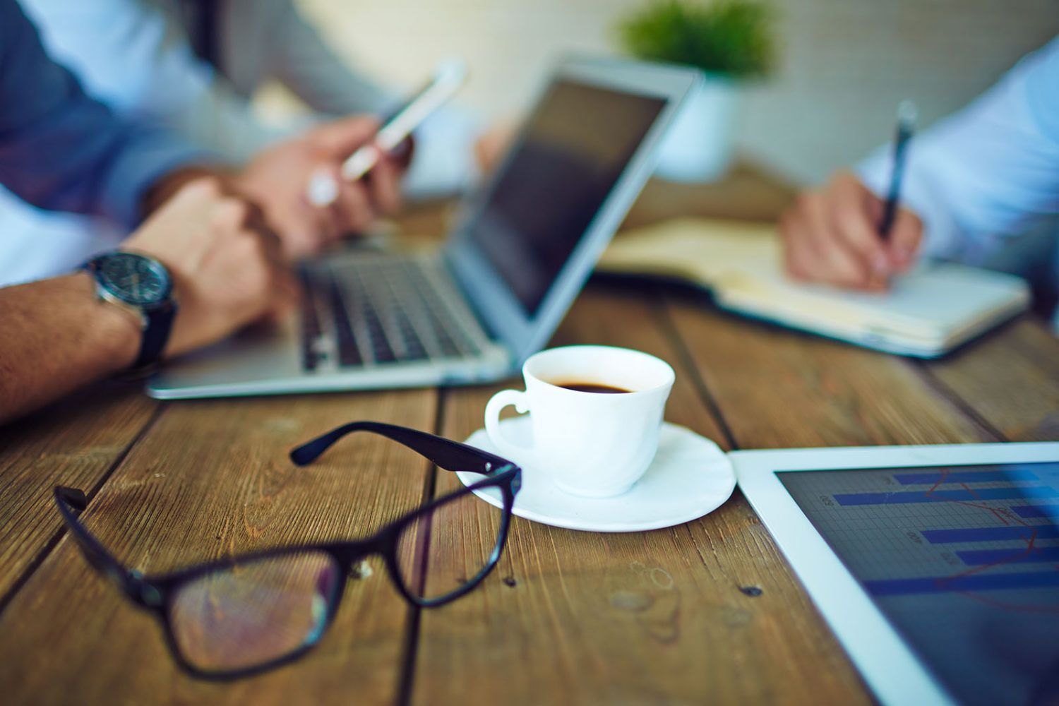 wooden table with eyeglasses and a white cup and saucer. men working on tablet, laptop and a man holding a cell phone.