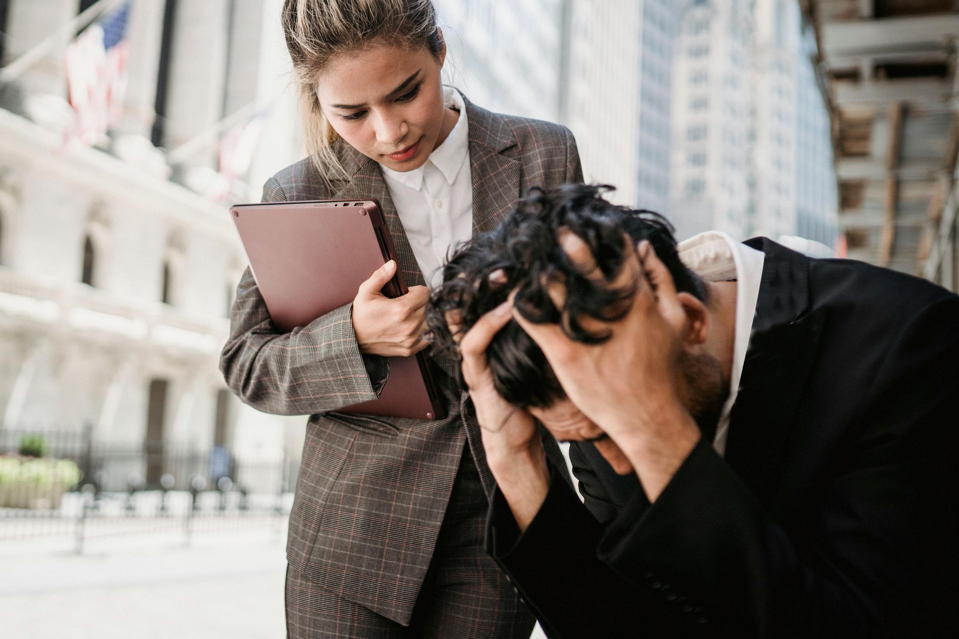 young woman in suit with laptop looking concerned over man in suit sitting with hands in hair seemingly not okay.