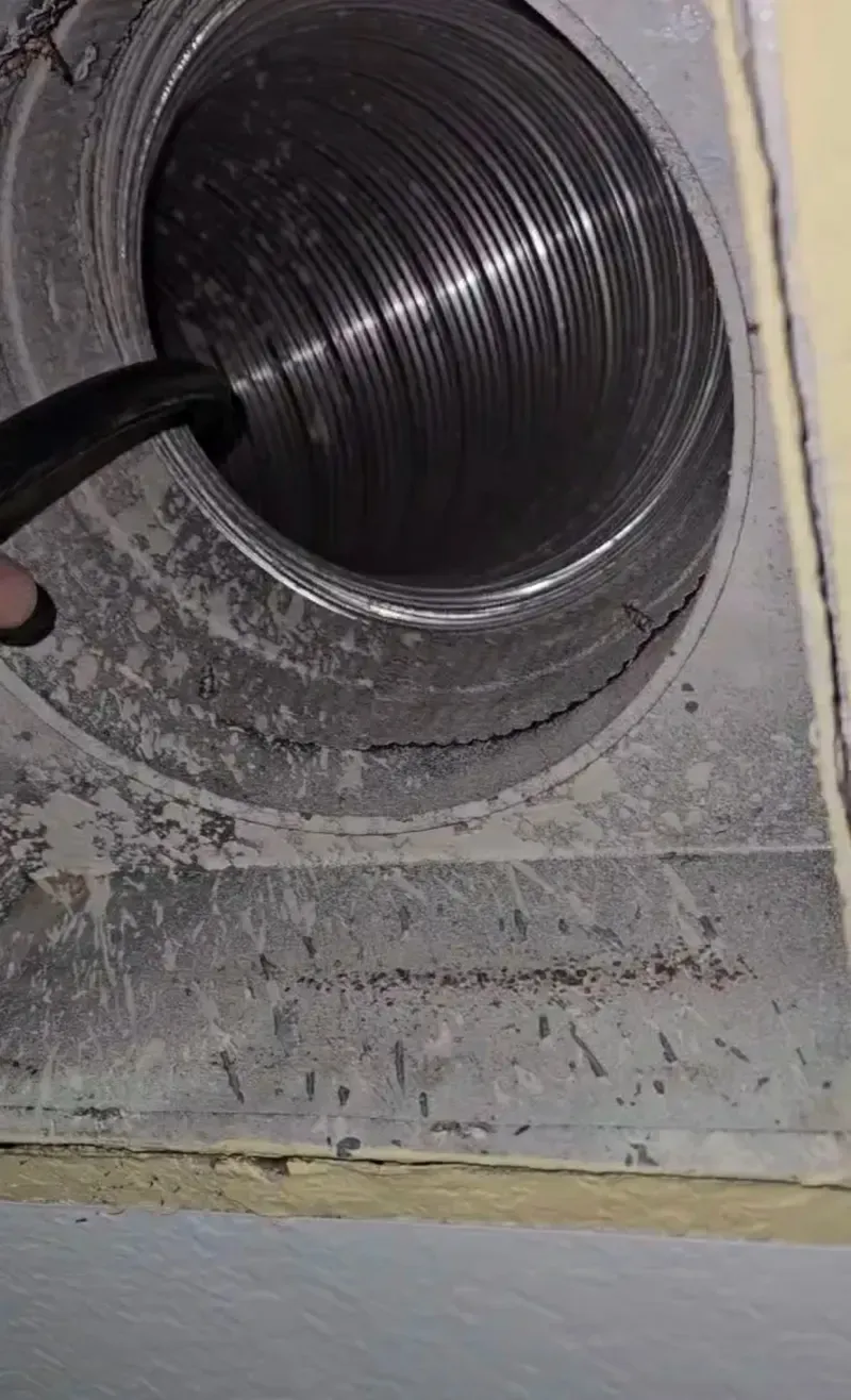 Round metal HVAC duct opening with a black hose inserted into a dusty wall panel.