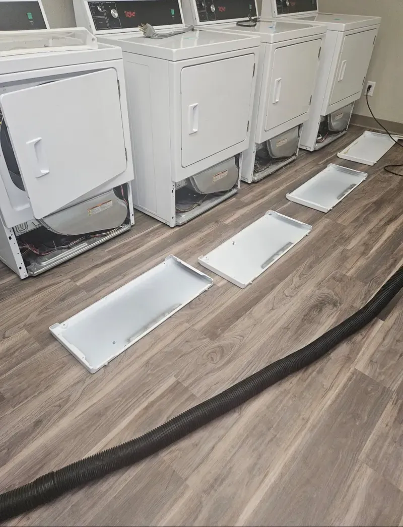 Dryer room with four white dryers and detached front panels laid on the wood floor.