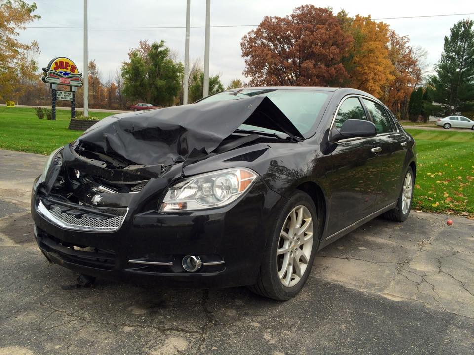 Black sedan with severe front-end damage, likely from a collision. It is parked near a sign and trees.